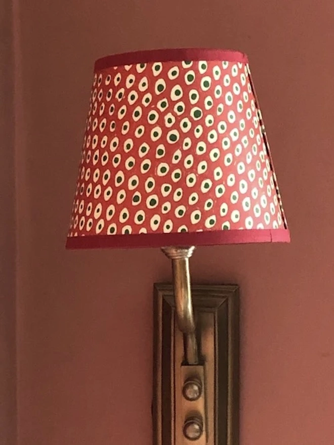 Candle Clip Shades. A red candle lampshade with cream and green spots and a crimson trim, fitted to a brass wall light against a dusky pink wall. The cheerful block-printed shade adds colour and charm to a bedroom or dining room.
