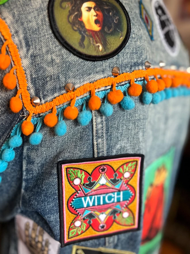 The patch has been embroidered onto a denim jacket, under an orange and blue trim detail. At the centre of the patch is the word “Witch” embroidered in white on a light blue strip. Surrounding this is a colourful, bold design which sits on a yellow background, with a thin pink inner border and a thick black outer border.