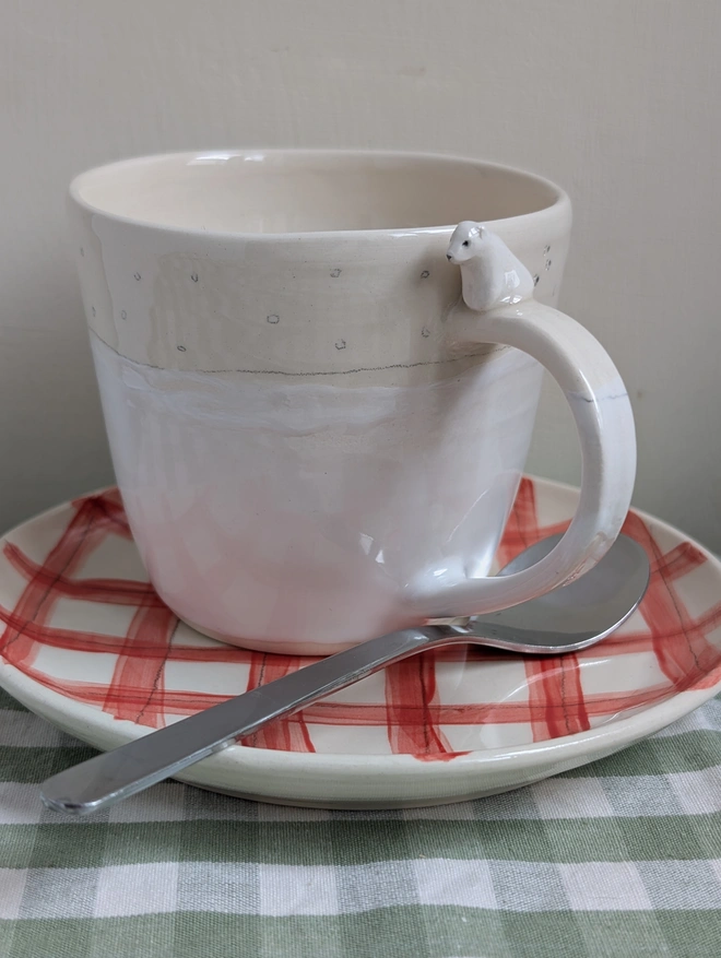 white cup with a ceramic polar bear sitting on the handle with tiny paw prints on the cup sitting on a red gingham plate