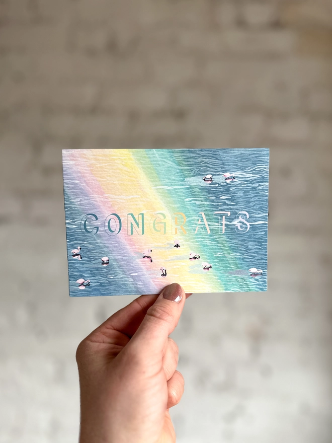 a greetings card featuring an illustration of of flamingos in a lake with a rainbow reflection with the text “CONGRATS”