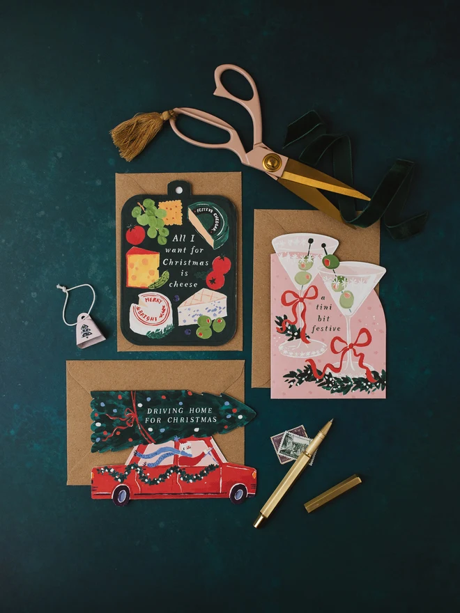 Cheeseboard Christmas card on a dark teal background with the Tini Bit Festive and Driving Home For Christmas cards, gold scissors and a gold pen