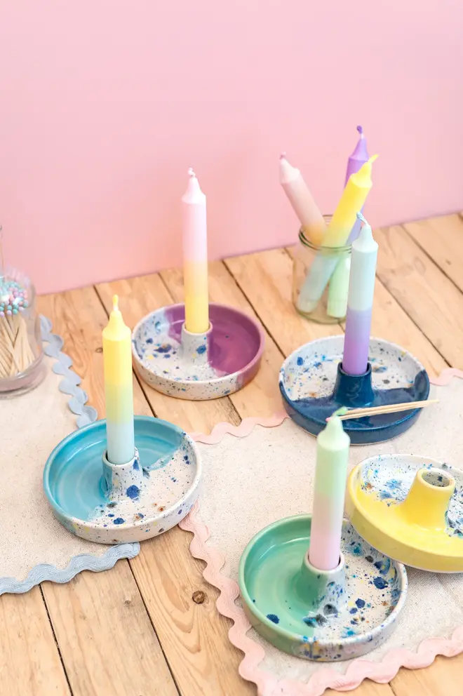 Shoreline Candle Stick Holder, a collection of colourful candle stick holders sitting on a wooden surface against a pink backdrop. They are accompanied by colourful napkins. 