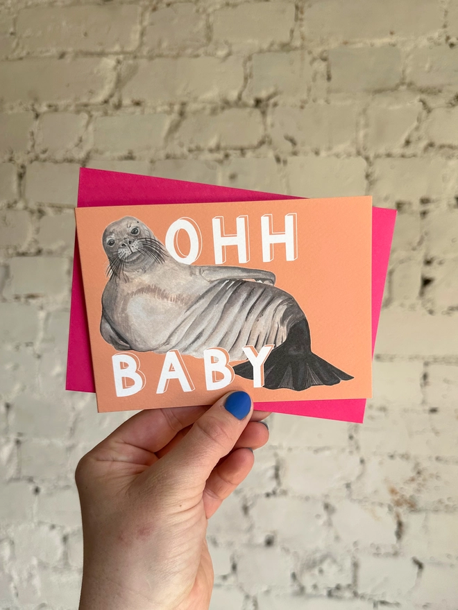 a peachy orange greetings card featuring an illustration of a chunky seal  with the text “ohh baby” with a neon pink envelope