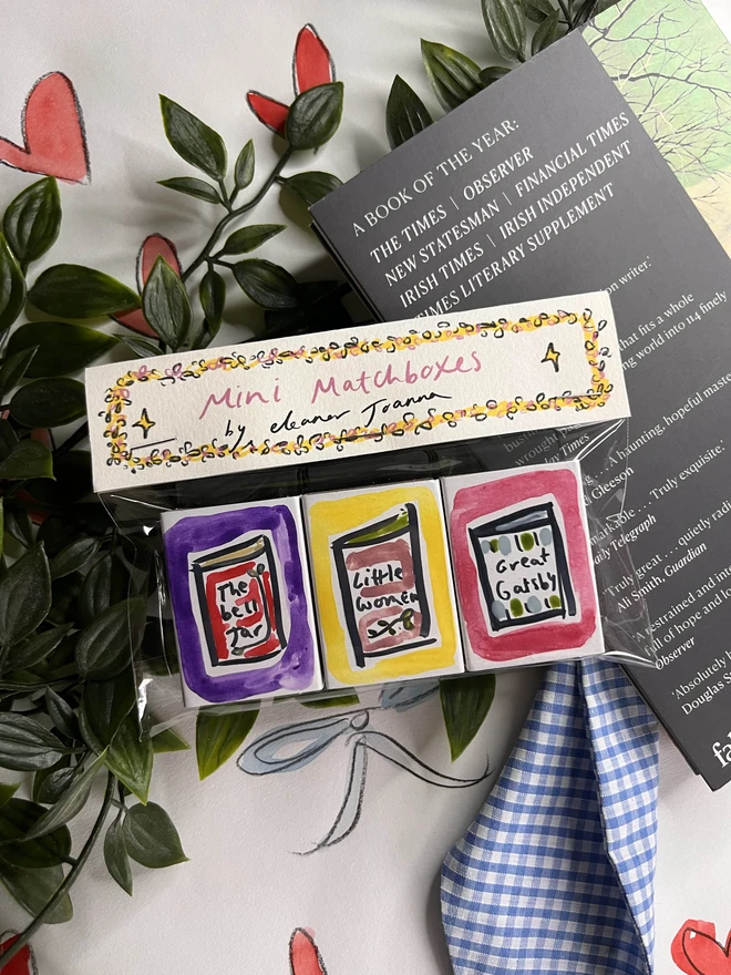 A collection of three hand-painted matchboxes with playful and colorful designs: one features two glasses with the word "Cheers!" written in bold lettering, another displays bright yellow lemons with "Easy Squeezy" written alongside, and the third showcases a red bow with the phrase "Bows & Arrows." Each matchbox is crafted with vibrant illustrations and creative lettering.