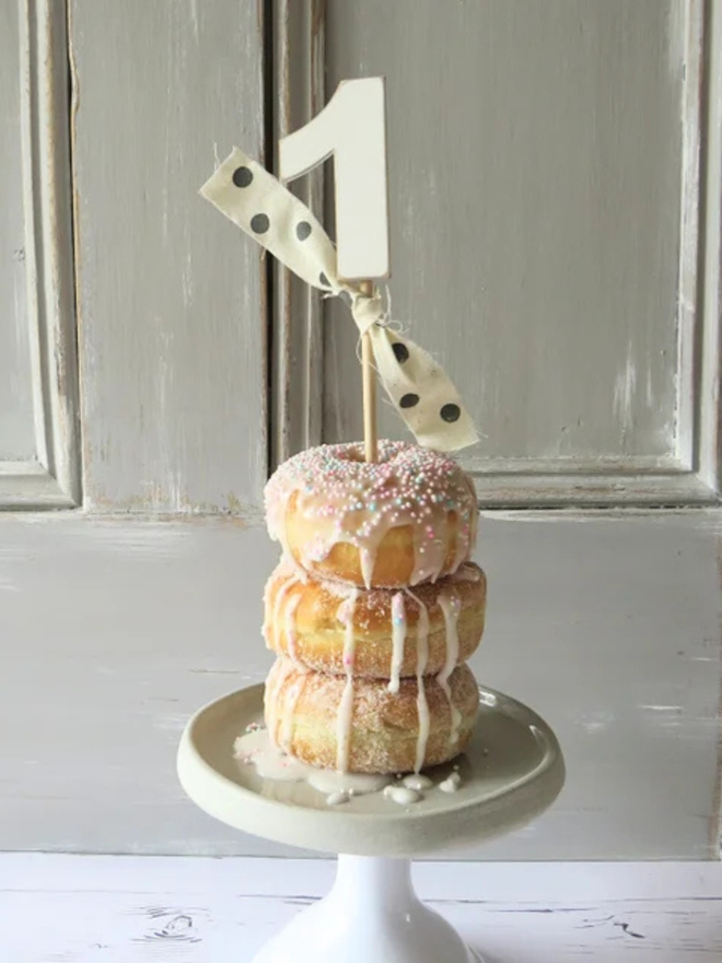 hand painted wooden number cake topper. a white number one cake topper with a spotty ribbon on a stack of donuts.