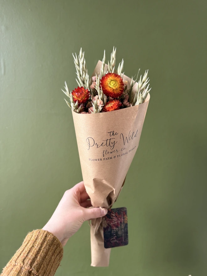 a bunch of dried flowers in bright colours wrapped in black tissue and kraft paper held by a person in a mustard coloured jumper with their left arm against a green wall