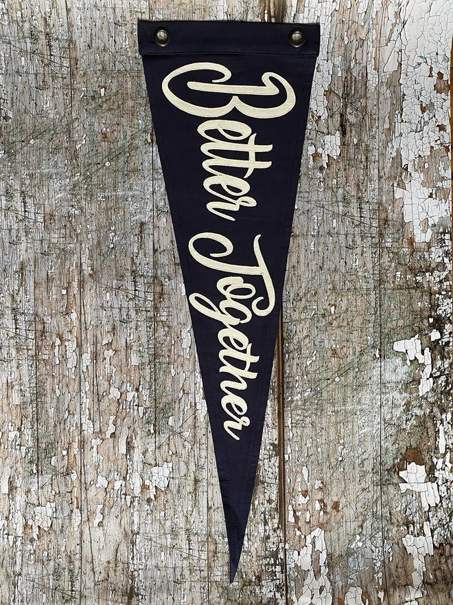 A navy canvas pennant flag hung vertically on a wooden wall. It has the words 'Better Together' written in ivory canvas.