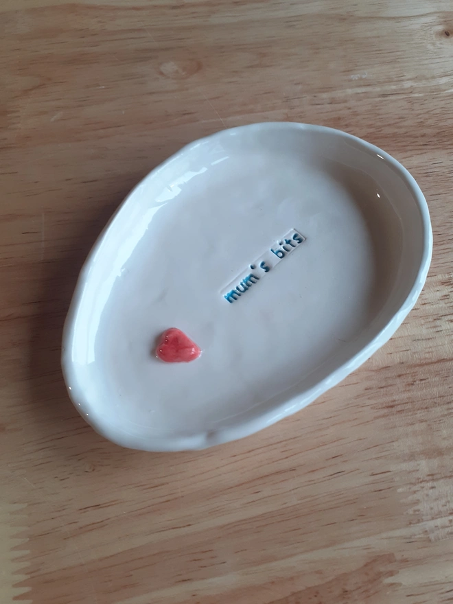 Handmade ceramic trinket dish on a wooden table, stamped with “mum’s bits” and finished with a small red heart, featuring slightly wonky oval shapes and glossy white glaze.