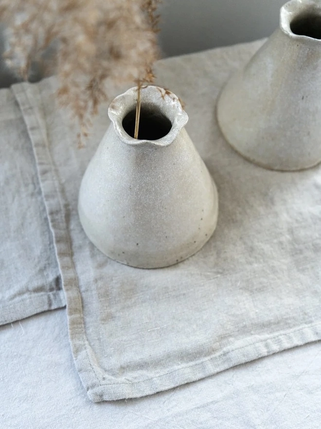 Maia Bud Vase In Sea Salt Glaze, with plant in it, placed on a table cloth against a grey background. 