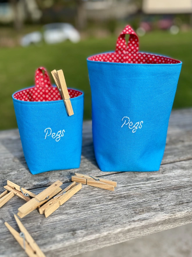Two blue cotton peg bags, one small, one large, embroidered with 'Pegs', on a garden bench.