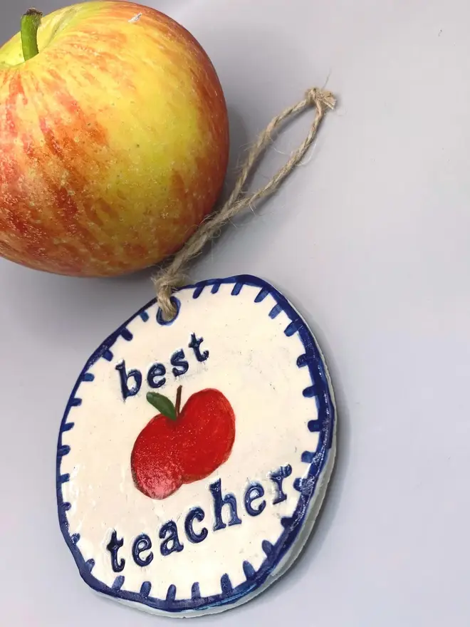 'Best Teacher' Handcrafted Ceramic Ornament, a ceramic apple ornament sitting on a plain white surface joined by an apple. 