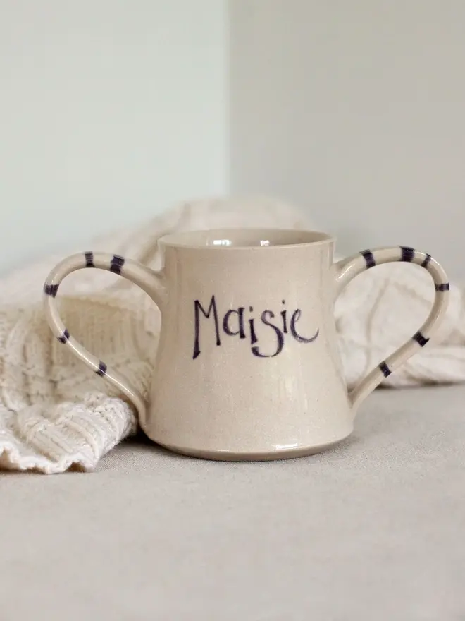 personalised double-handled ceramic mug. mug emblazoned with name 'maisie' and photographed on white shelf. 