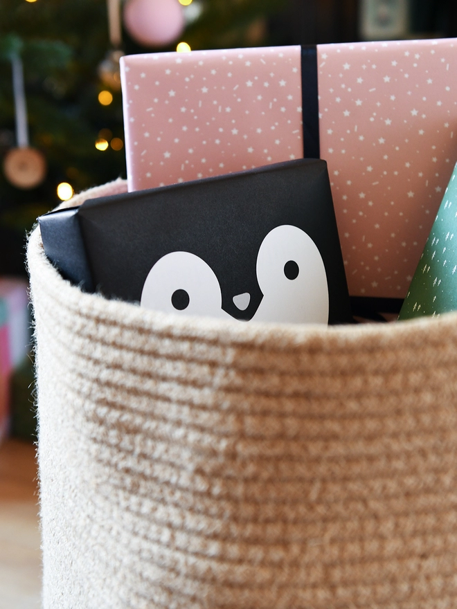 A gift wrapped in penguin wrapping paper is tucked into a woven basket with several other wrapped gifts.