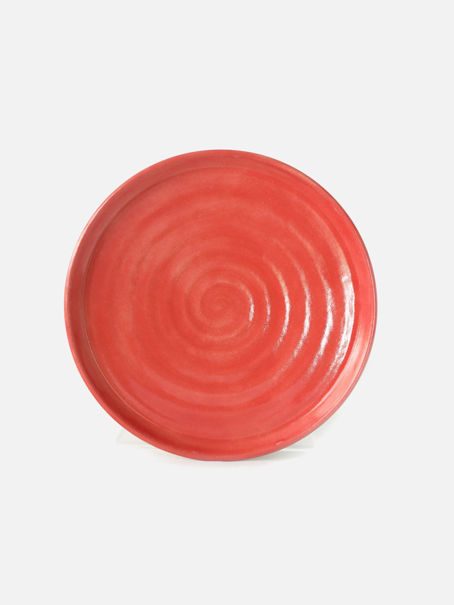 A handmade ceramic cake plate in cherry red glaze, photographed from the front against a plain light grey background, showing a natural concentric swirl texture and a shallow raised rim