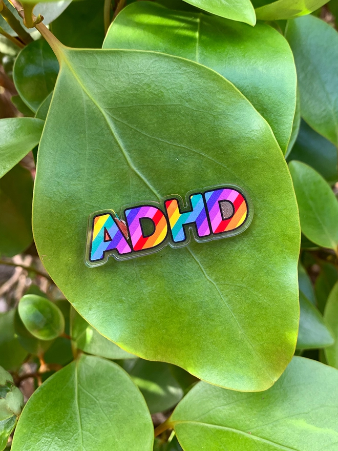 image shows an acrylic badge in the shape of the letters ADHD. the letters are filled with diagonal rainbow stripes.