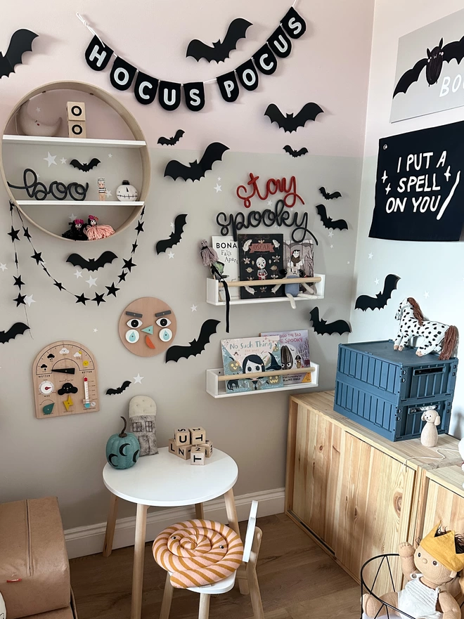 'stay spooky' wire word signs used as Halloween decorations in a Scandi Style Kid's playroom or bedroom. 'stay spooky' is hanging above two bookshelves on the wall with Hallow's eve Books and Halloween Props decorating the space. There are more signs and children's toys are sitting on a circular shelf in this playroom surrounded by lots of kid's toys and play room furniture.