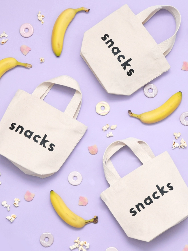 three mini kid's size tote bags with the word snacks on it laying on a purple backdrop with bananas sweets and biscuits surrounding them