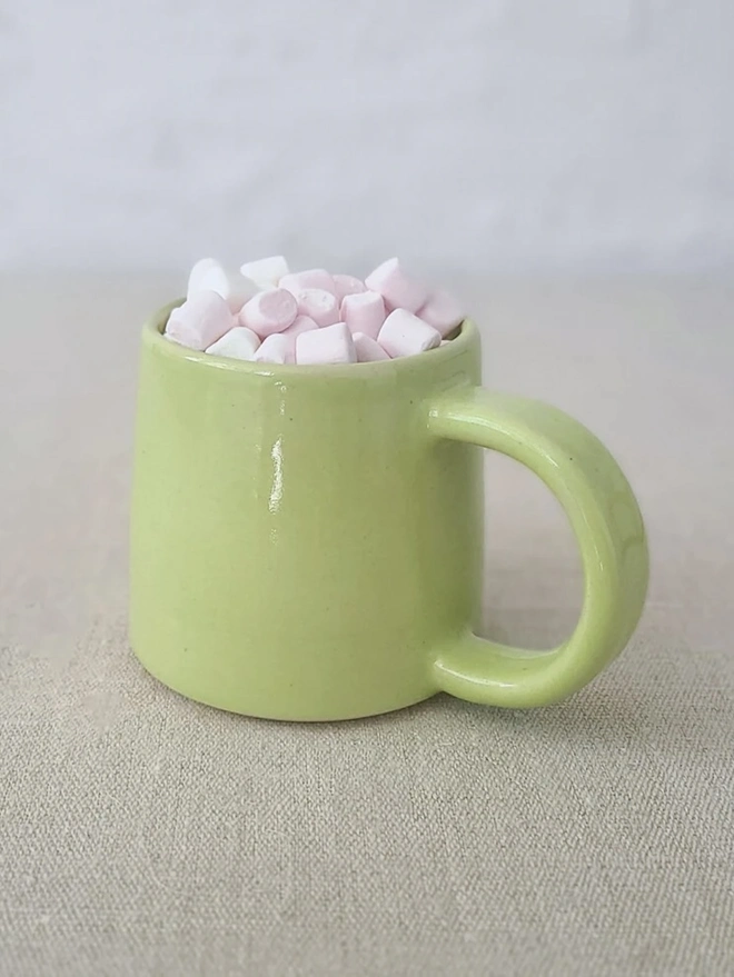 Ceramic Standard Mug, a colourful ceramic mug sitting on a plain surface against a grey backdrop. It is accompanied by marshmallows. 
