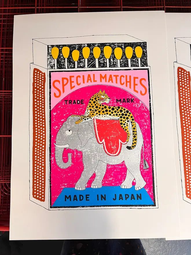 Elephant And Leopard Matchbox Taken For An Extra Long Rid, a colourful matchbox design in the centre of a white card. It sits on a studio rack. 