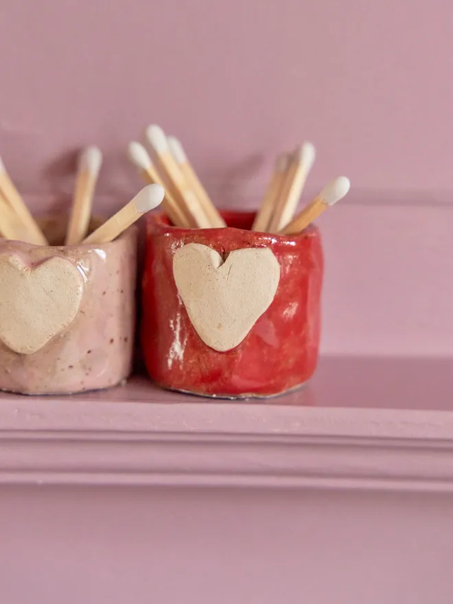 Mini ceramic match holder in red and pink on a pink shelf with white matches inside.