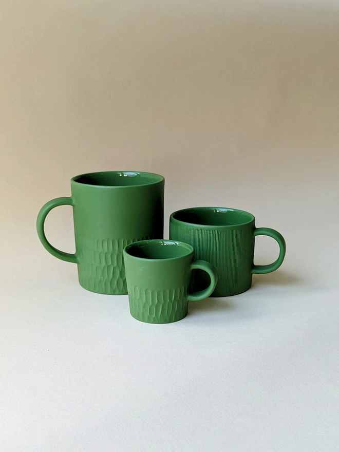 Three handmade porcelain mugs and coffee cups in dark green. A large mug for tea with a hand carved texture around the base, a small mug for flat white coffee which is hand carved all over, and an espresso cup with hand carving around the base. All are glazed inside and matte outside.