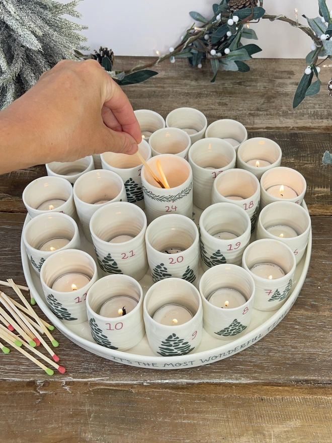 alternative advent calendar made up of a gorgeous circular porcelain tray with 24 individual porcelain cylinders for each day of advent. hand lighting the 24th candle holder while it sits on a rustic table with christmas tree in background