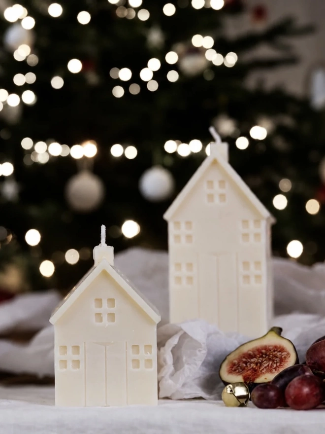 White Christmas Townhouse Candles (Set Of 3). Two cream house candles in different sizes, with detailed windows and doors, styled on linen with figs, grapes and a gold bell before a twinkling Christmas tree. Together they create a sweet miniature village scene for the mantelpiece or table.