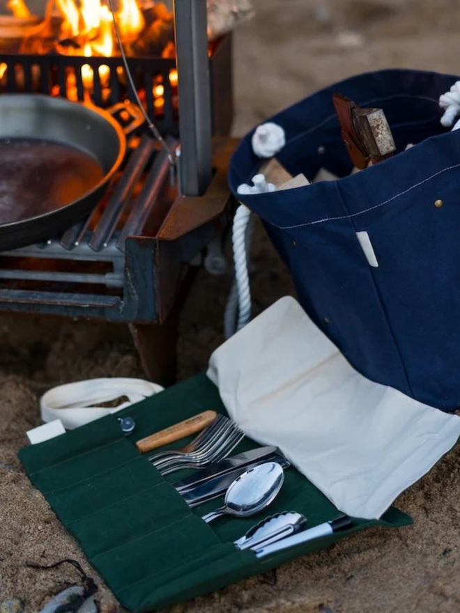 'mess kit' cotton cutlery and utensil roll. a pack of picnic utensils in a cotton roll, open and photographed next to a camp cooking setup.