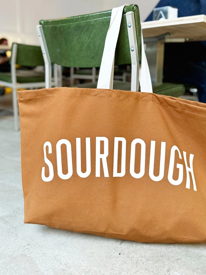oversized tan canvas tote bag with sourdough slogan hanging on the back of a green chair