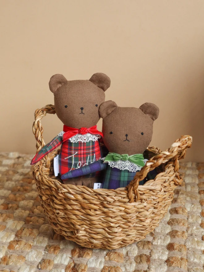 Two handmade linen teddy bears wearing festive tartan outfits, one in red and green with embroidered “Teddy” lettering and a red bow, and the other in green and navy tartan with a green bow. Both feature delicate lace collars and embroidered faces.