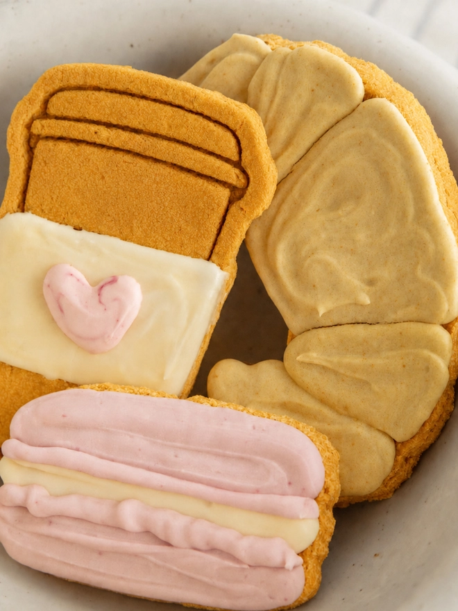 Close-up of croissant, takeaway coffee cup and pink macaron Parisian Bakery iced dog biscuits arranged in a bowl.