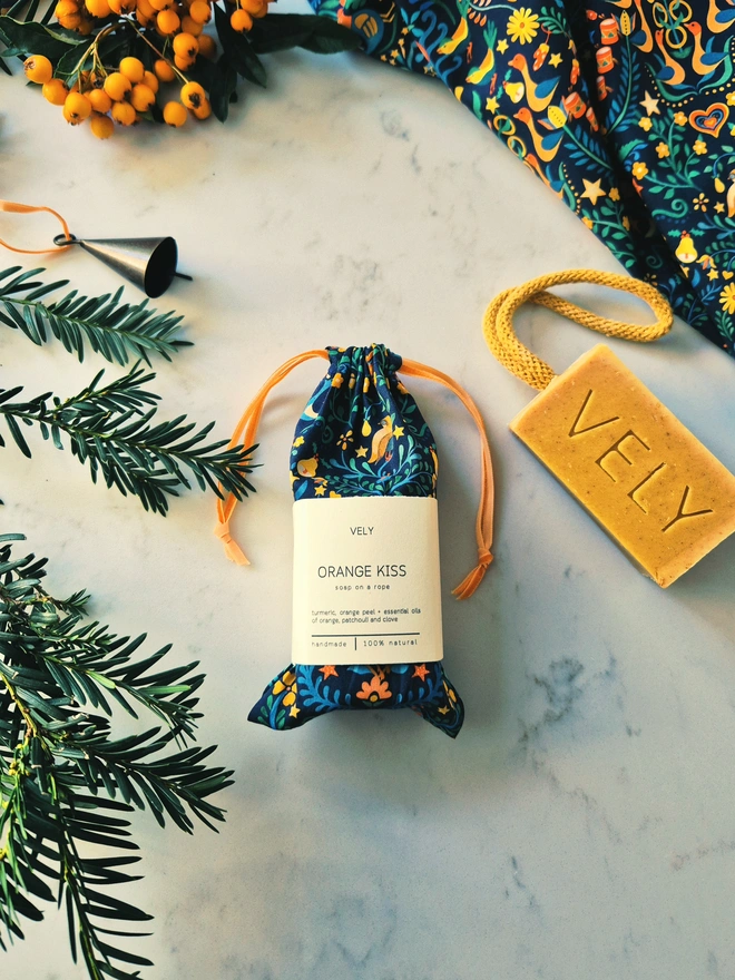 Orange Kiss soap on a rope in a festive Liberty print bag, displayed beside an evergreen branch.