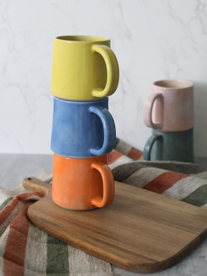 Three handmade ceramic standard mugs stacked on a wooden board in yellow, blue, and mandarin orange, with blush pink and teal mugs softly blurred behind, styled with a striped linen cloth against a white marble background