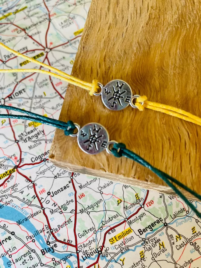 Adventure wish bracelets with a silver compass charm laying on a piece of wood with a map.