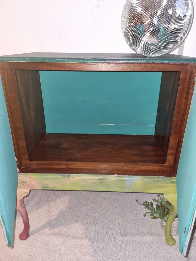 A painted record cabinet decorated with a sci fi image from the 1950s of a lady running from an alien and on the other door, a rose taken from a Victorian seed packet. There is wording that runs down the middle of the cabinet which reads 'it could be worse'. The colours are apple green and pinks.