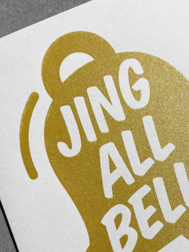  Close up view of a handprinted christmas card, sparkly gold ink in the shape of a bell with the words Jing All Bells within.