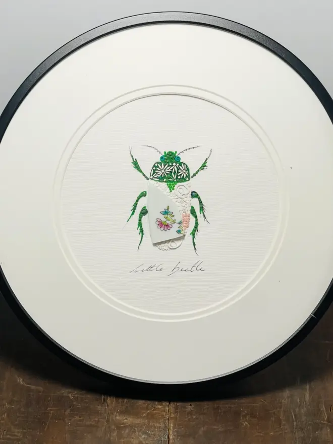 Hand-drawn, painted, mosaic clay, vintage floral pottery insect on white double mounted card, in a black circular frame.
