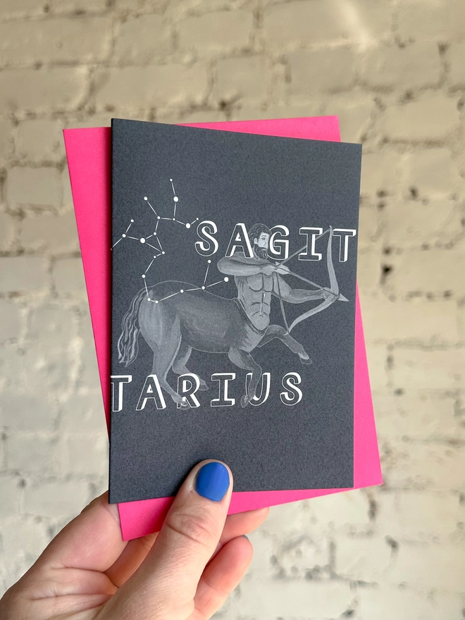 a greetings card featuring a greyscale illustration of a zodiac style centaur with the text “SAGITARIUS” with a neon pink envelope