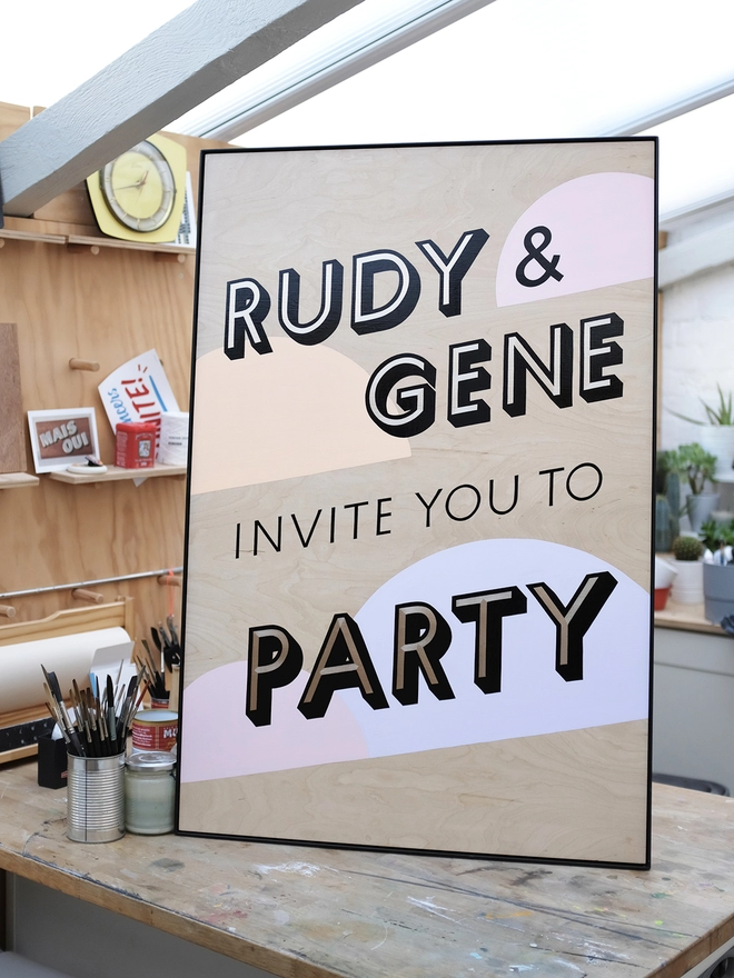 Handpainted invite to party sign with gold leaf letters outlined in black, with pastel coloured semicircles in the background. Shown in the artist's workshop. 