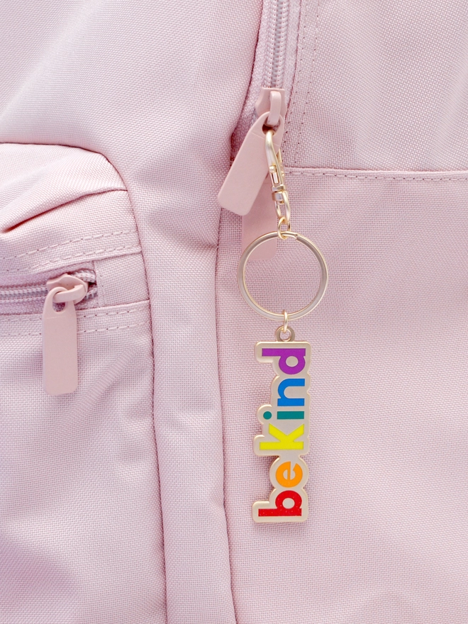 Enamel keyring with 'Be Kind' design in rainbow colours hanging from a pink backpack