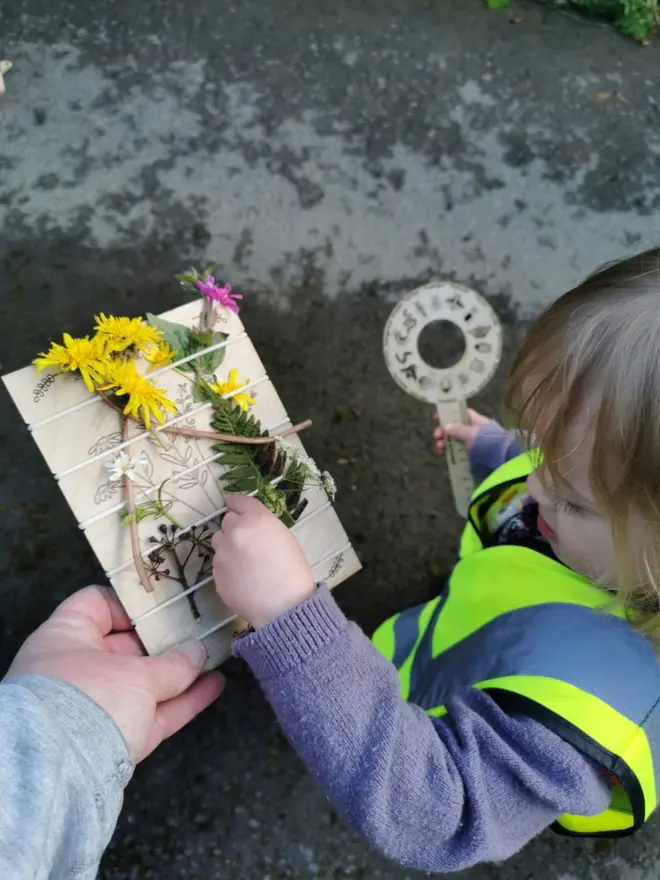 Birch wood nature trail weaving board decorated with artificial wildflowers and leaves, held in place by cream-colored cord. Toddler is placing a flower. 