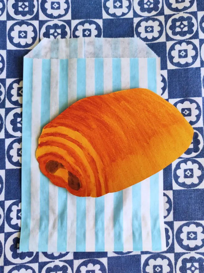 hand painted pain au chocolat, cut out paper shape. with blue and white striped paper bag