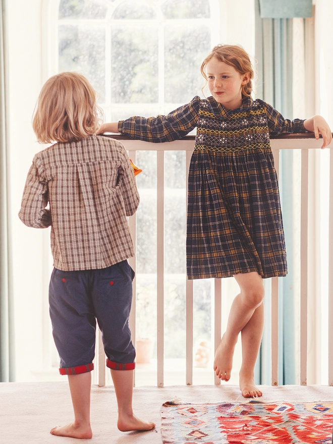 A girl hangs off railings wearing a navy and yellow dress with hand smocked detail talking to a boy in a plaid shirt and cropped trousers