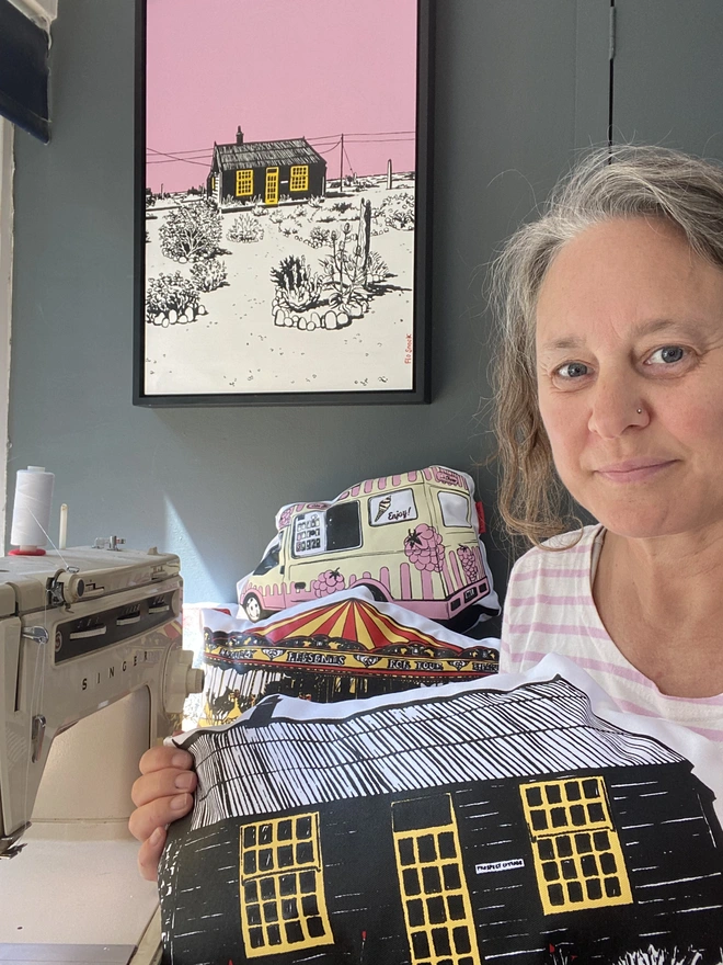 A prospect cottage cushion, handmade by artist Flo Snook. The artist holding the cushion in her studio, with her singer sewing machine alongside and her painting on the wall in the background