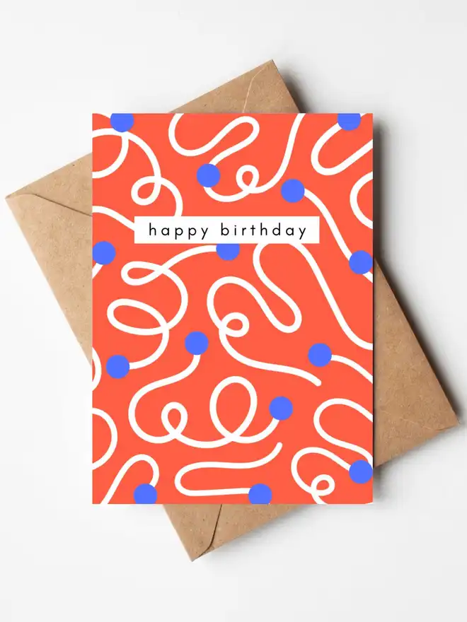 A modern abstract birthday card featuring flowing white lines and blue dots on a bright coral background, with the words "happy birthday" across the centre. The card is displayed with a kraft envelope.