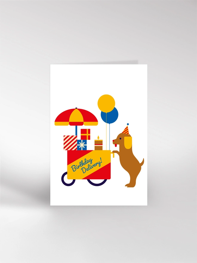 Birthday card with a dog pushing a wagon laden with gifts, balloons and cake