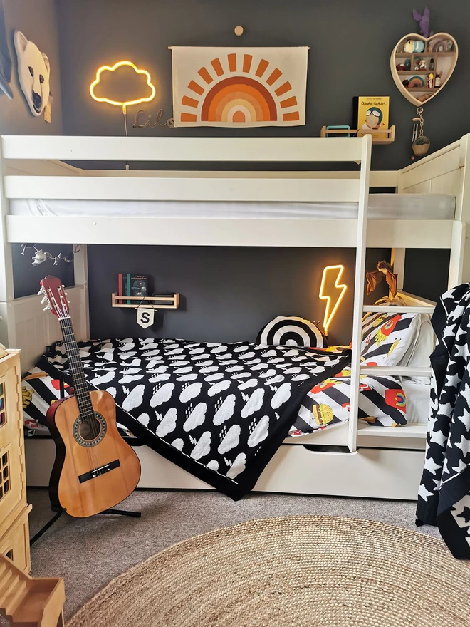 A view of white single bunk beds in a children's room, a black and white blanket with a storm cloud pattern is draped at an angle over the bottom bunk. Above the bed hangs a fabric banner with an orange sunset, and cloud and lightning bolt neon wall lights are switched on creating a warm and cosy feel. A guitar is propped on a stand near the side of the bed, and in the foreground a wooden dolls house is just visible.