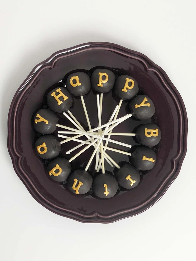 Black And Gold 'Happy Birthday' Cake Pops, cake pops placed onto a black plate. This is against a white backdrop. 
