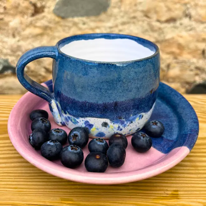 Shoreline Large Round Mug, a colourful mug on a wooden surface against a brick backdrop. It is accompanied by fruits. 