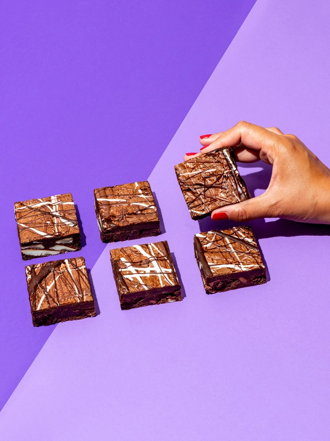 Triple Chocolate Brownies (Box Of 6), handing picking up a brownie against a purple backdrop. 
